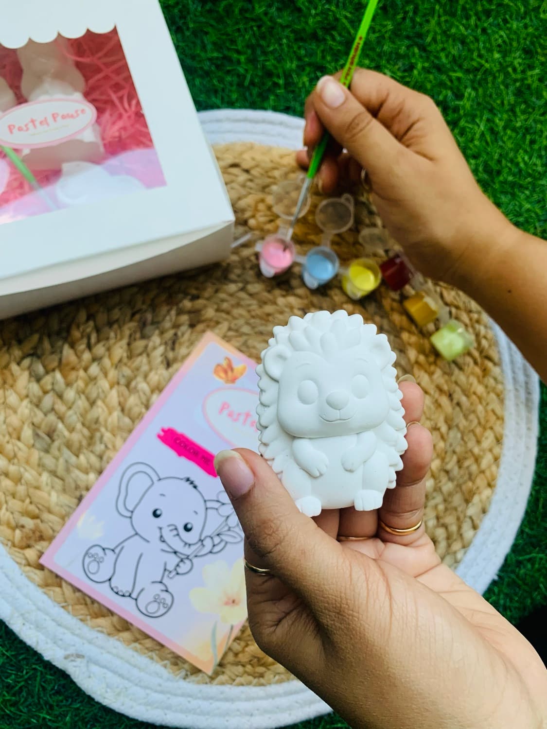 A hand holding an unpainted white hedgehog figurine beside a Pastel Pause kit, Color Me card and pastel paint pots